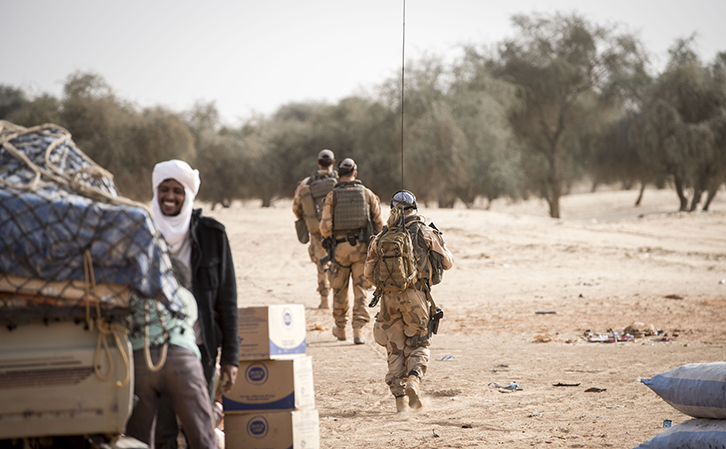 Tre soldater med blå baskrar går längs en dammig väg med träd intill. I förgrunden syns lådor och packning samt en civil person.