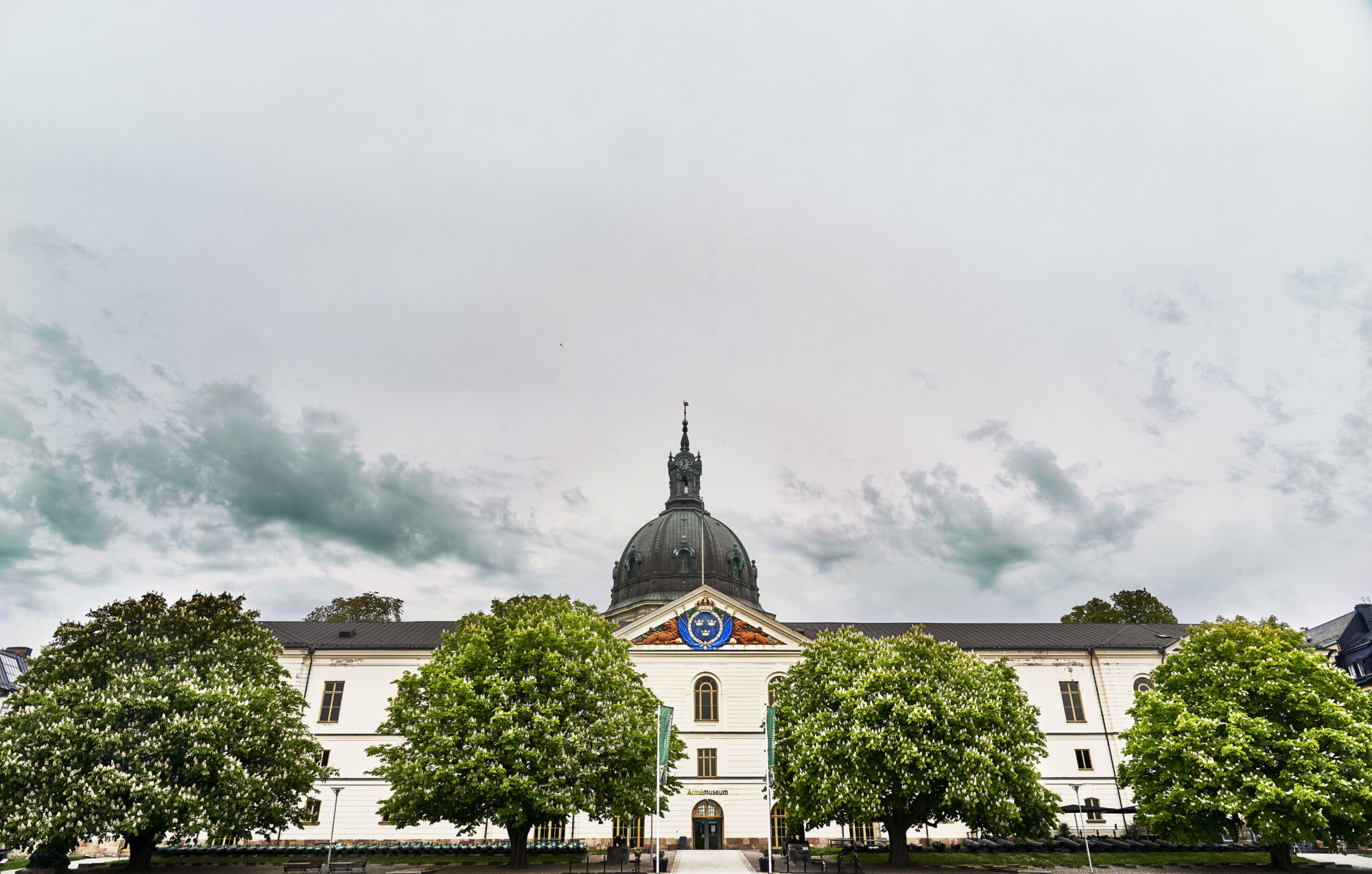 Vit byggnad med svart kupoltak över entrén, sedd från gatan omgiven av gröna träd under en molnig himmel.
