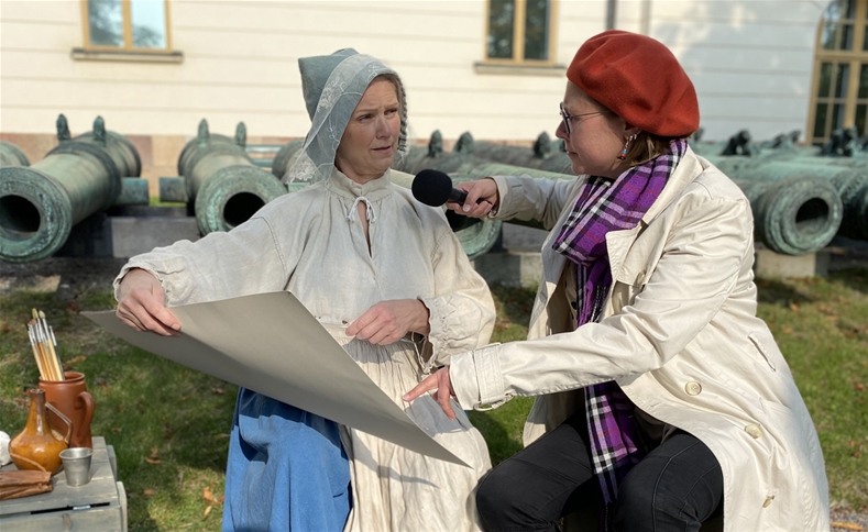 En person i röd basker och ljus rock håller en mikrofon framför en person klädd i äldre kläder.