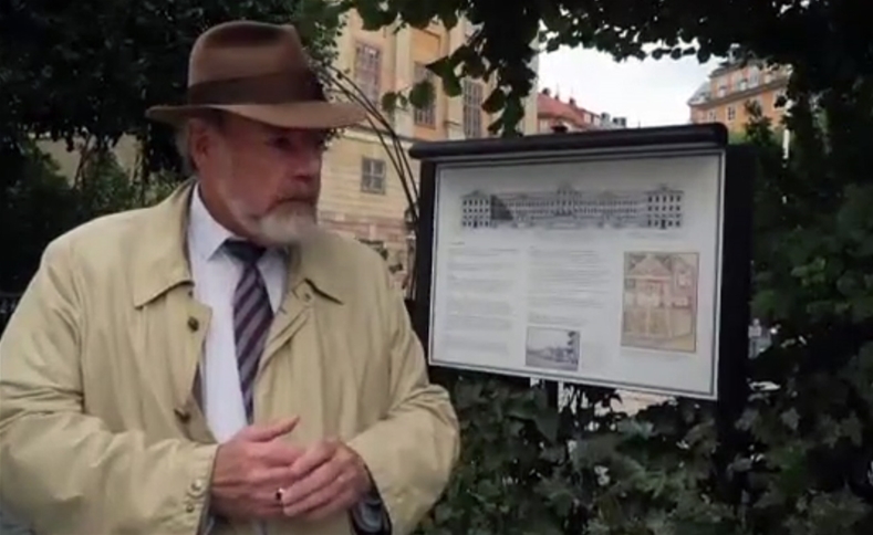 En person i rock hatt står vid en informationsskylt med grönska omkring, personen är Thomas Roth som håller en stadsvandring på Östermalm.