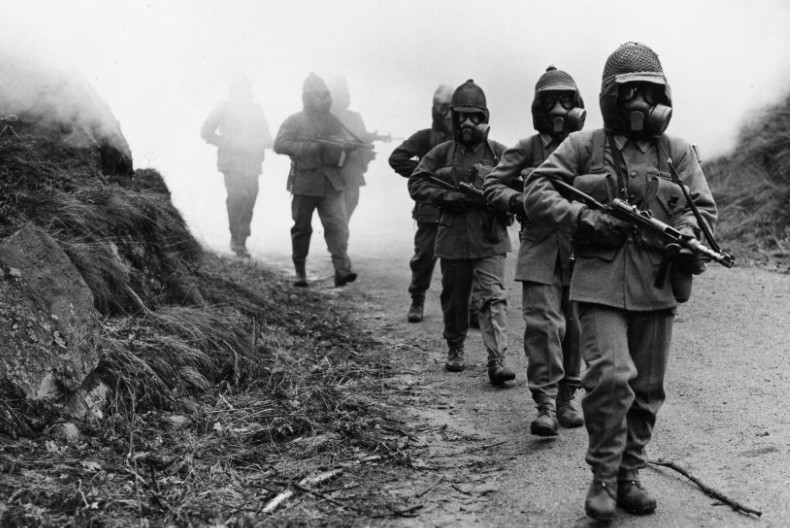 Soldater går på led med vapen i famnen och gasmask över ansiktena.