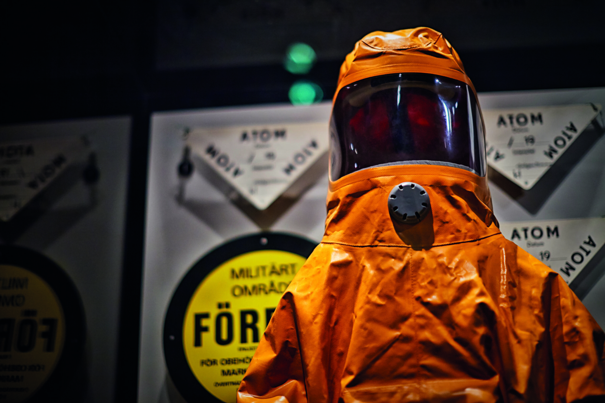 En helkroppstäckande orange skyddsdräkt framför uppsatta varningsskyltar som det bland annat står "Atom" och "Militärt område" på.