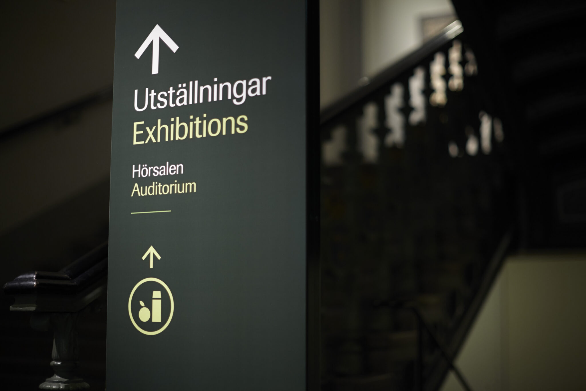 En grön skylt som det står "Utställningar" på samt en pil som pekar uppåt. I bakgrunden syns en trappa upp till nästa våning.