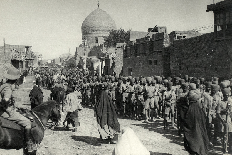 Svartvit bild av militärparad på en stadsgata, med rader av soldater i turbaner och korta uniformer. I bakgrunden syns en byggnad med kupoltak.