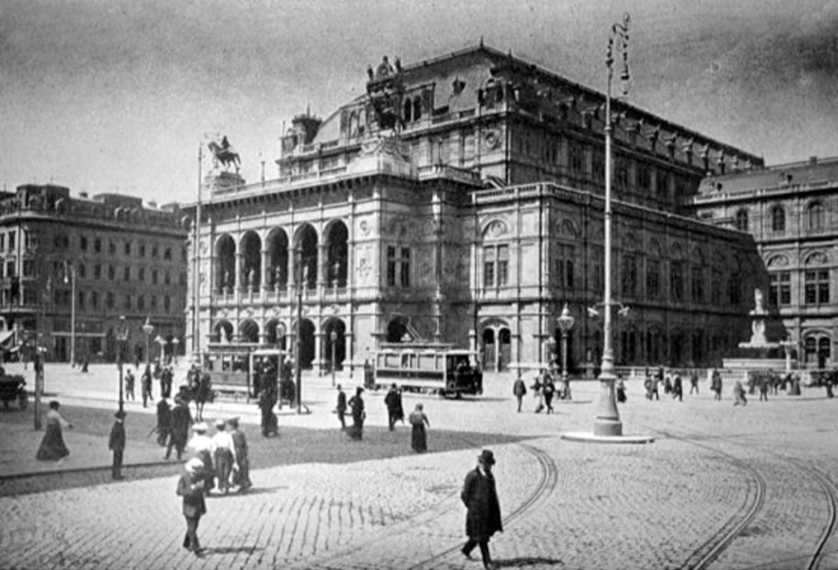 En stor pampig byggnad, Wiens statsopera. På det stenbelagda torget framför operan står två äldre spårvagnar och människor promenerar omkring.