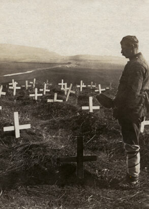 En man med mössan i hand står vid ett av många kors nedstuckna i jorden på en stor äng.