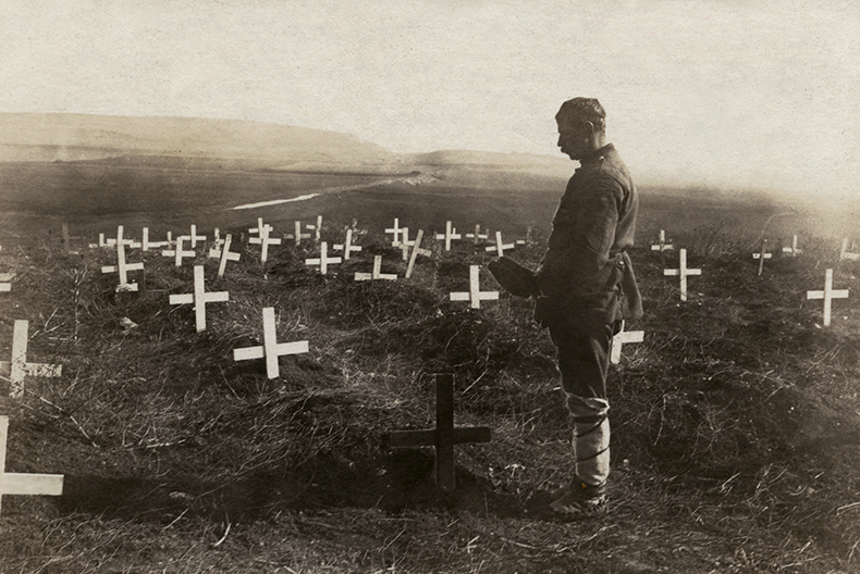 En man med mössan i hand står vid ett av många kors nedstuckna i jorden på en stor äng.