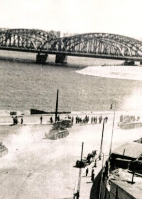 Militärfordon kör längs vatten. Längre bort syns en lång bro.