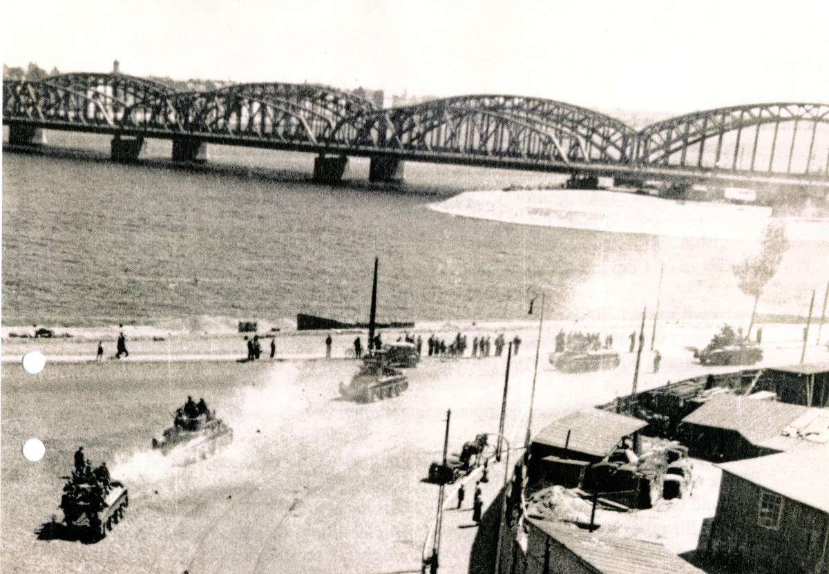 Militärfordon kör längs vatten. Längre bort syns en lång bro.