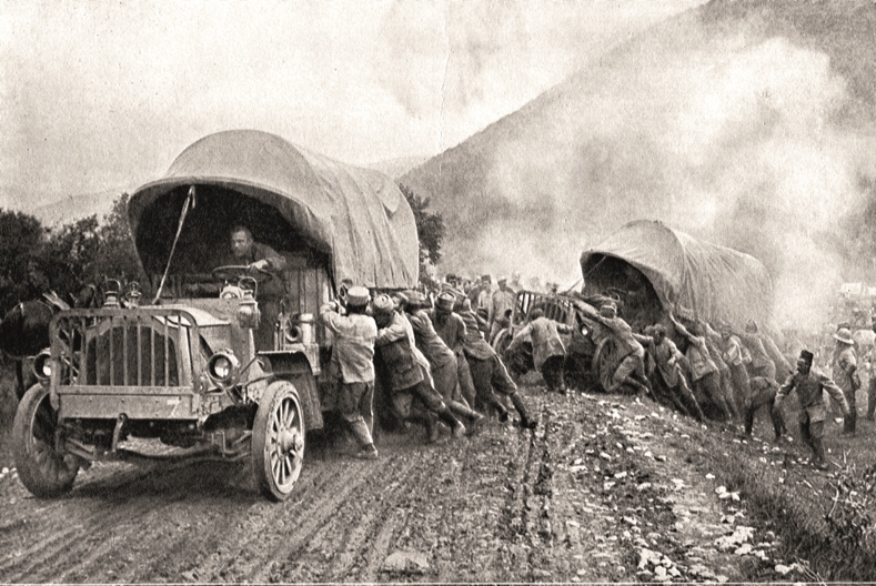 Svartvit bild av en stor folkmassa som hjälper två lastbilar upp på vägen igen. Vägen är lerig och den ena lastbilen verkar ha problem att komma upp ur ett dike.