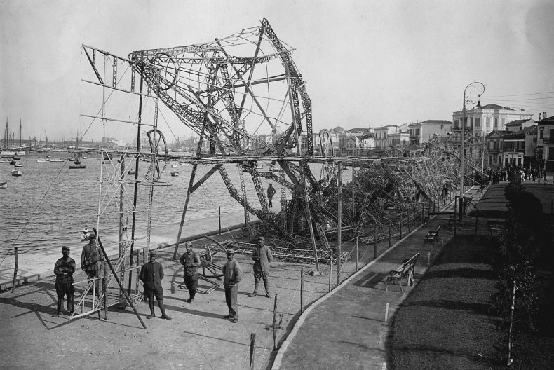En grupp personer står framför något som mest påminner om rasade byggställningar invid vattnet i en hamn.