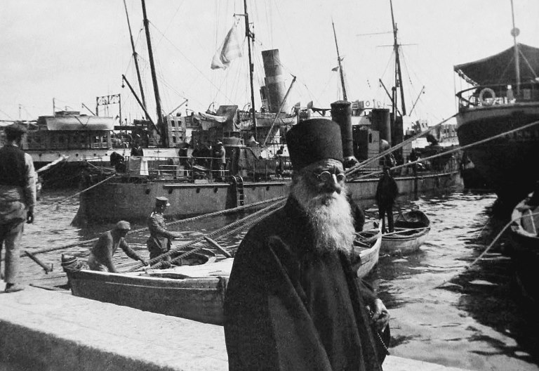 En person med långt skägg och cylinderformad hatt står i en hamn och bakom syns både små och större båtar med besättning.