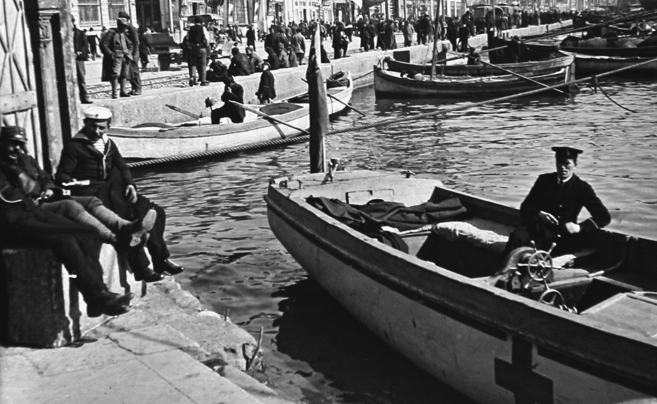 Svartvit bild av flera små båtar vid en kaj, med människor som sitter och står längs vattnet.
