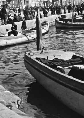 Svartvit bild av flera små båtar vid en kaj, med människor som sitter och står längs vattnet.