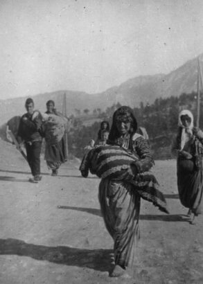 Svartvit bild av människor på flykt som går på en väg med skog och berg i bakgrunden. Längst fram en kvinna med ett stort bylte i famnen.