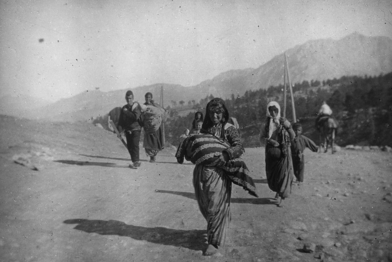 Svartvit bild av människor på flykt som går på en väg med skog och berg i bakgrunden. Längst fram en kvinna med ett stort bylte i famnen.