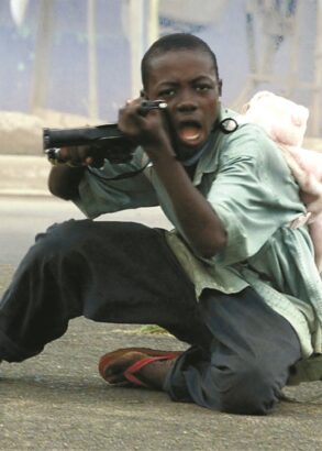 Ett barn med en ryggsäck i form av en nalle på ryggen riktar ett vapen mot den som tog bilden.