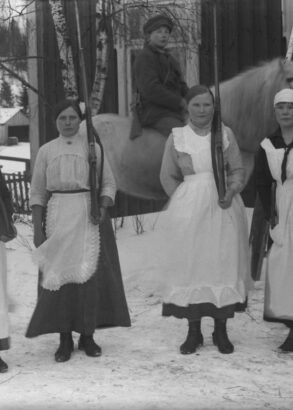Framför ett hus står fyra personer iförda klänning och vita förkläden på rad med varsitt gevär lutat mot vänster axel, bakom dem sitter en person till häst. Det är vinter.