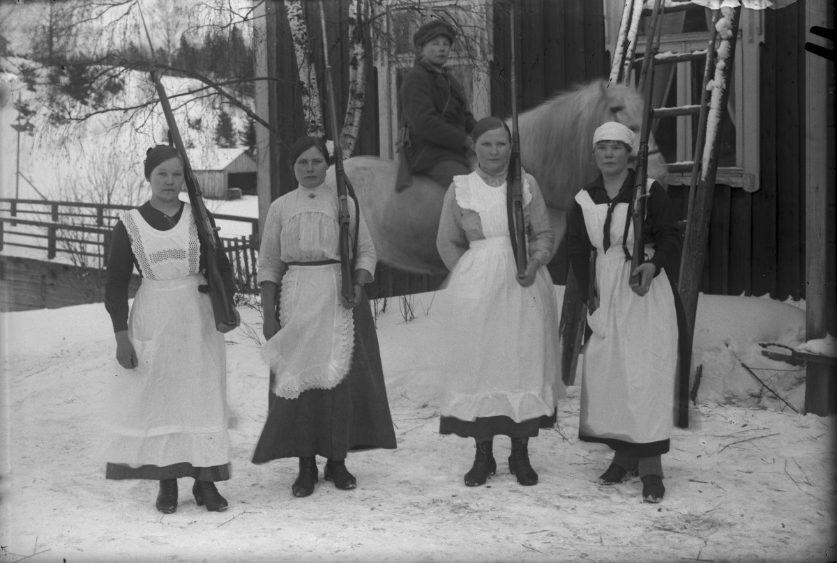 Framför ett hus står fyra personer iförda klänning och vita förkläden på rad med varsitt gevär lutat mot vänster axel, bakom dem sitter en person till häst. Det är vinter.