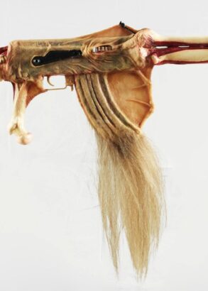 Ett konstnärligt vapenliknande föremål skapat av material som påminner om djurben, hud och hår. Objektet har formen av ett gevär men består av organiska former med en ryggradsliknande kolv, benformade detaljer och en lång tofs av ljust hår som hänger från undersidan. Bakgrunden är helt vit.