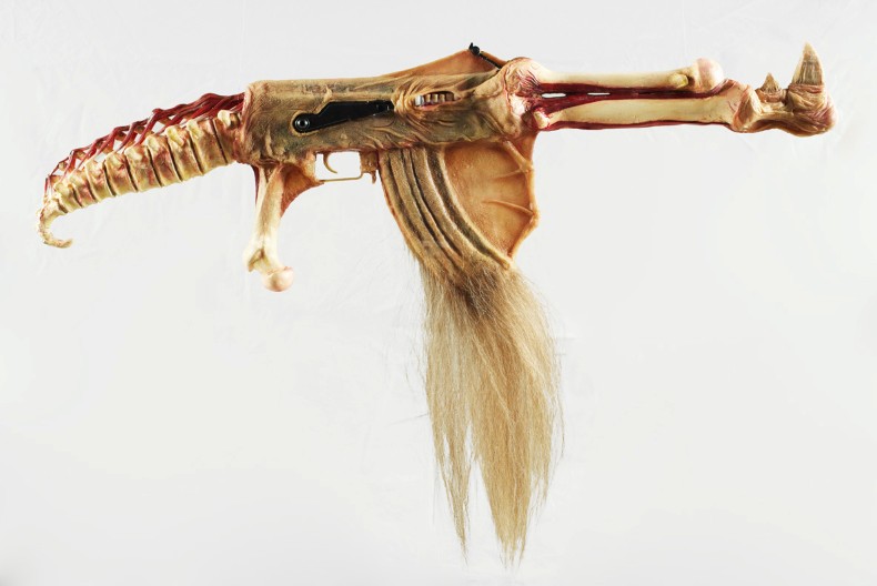 Ett konstnärligt vapenliknande föremål skapat av material som påminner om djurben, hud och hår. Objektet har formen av ett gevär men består av organiska former med en ryggradsliknande kolv, benformade detaljer och en lång tofs av ljust hår som hänger från undersidan. Bakgrunden är helt vit.