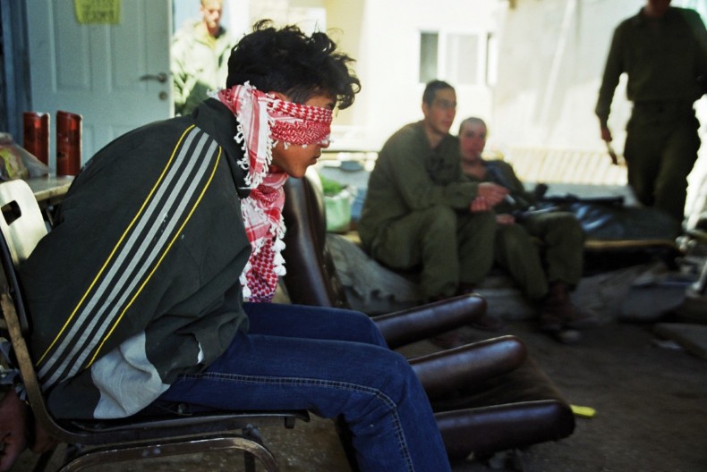 I förgrunden en bakbunden man på en stol och med förbundna ögon. I bakgrunden tre grönklädda män som ser ut att vänta på något.