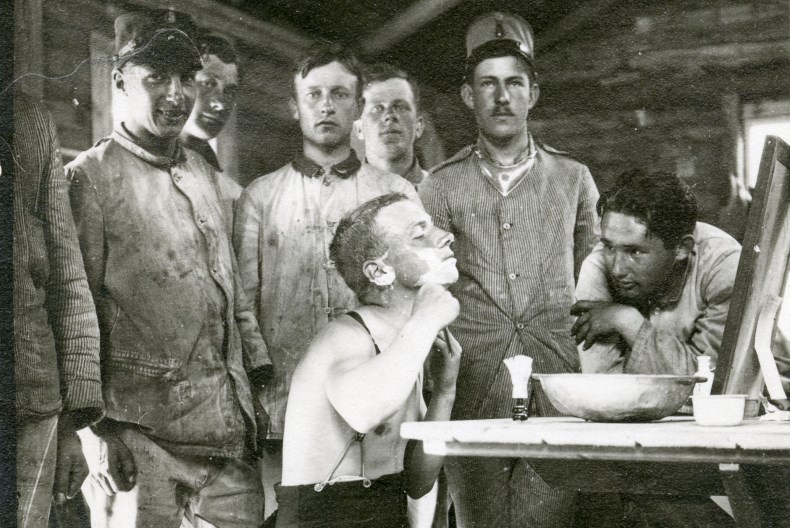 En person sitter framför spegeln vid ett bord med en vattenskål framför sig och rakar sig medan sju andra personer tittar på.