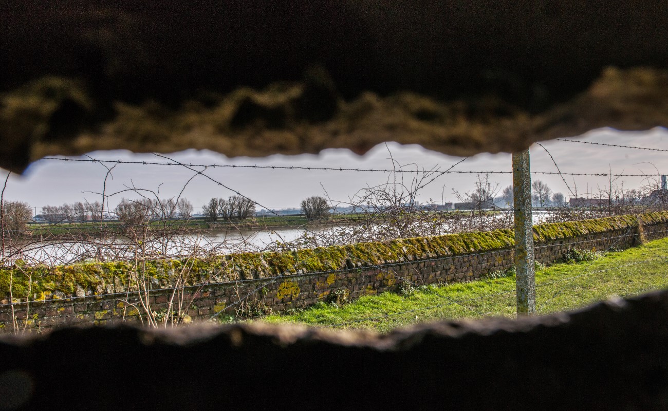 Som att titta ut ur ett gömställe och se en lång överväxt mur med taggtråd på som följer vattnet. På andra sidan vattnet skymtas träd och grönområden.