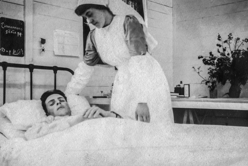 En sköterska i sköterskeuniform böjer sig ner över en skadad soldat nedbäddad i en säng med vita lakan.