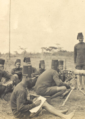 Svartvit bild av soldater i fält. flera sittandes på marken och några stående vid ett maskingevär med en officer i hjälm bredvid.