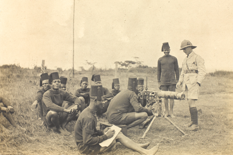Svartvit bild av soldater i fält. flera sittandes på marken och några stående vid ett maskingevär med en officer i hjälm bredvid.
