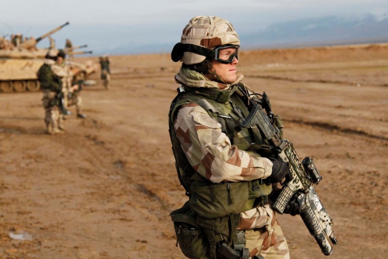 En soldat i full mundering och ett stort vapen i händerna står och blickar mot horisonten. Snett bakom soldaten står en stridsvagn och fler soldater.