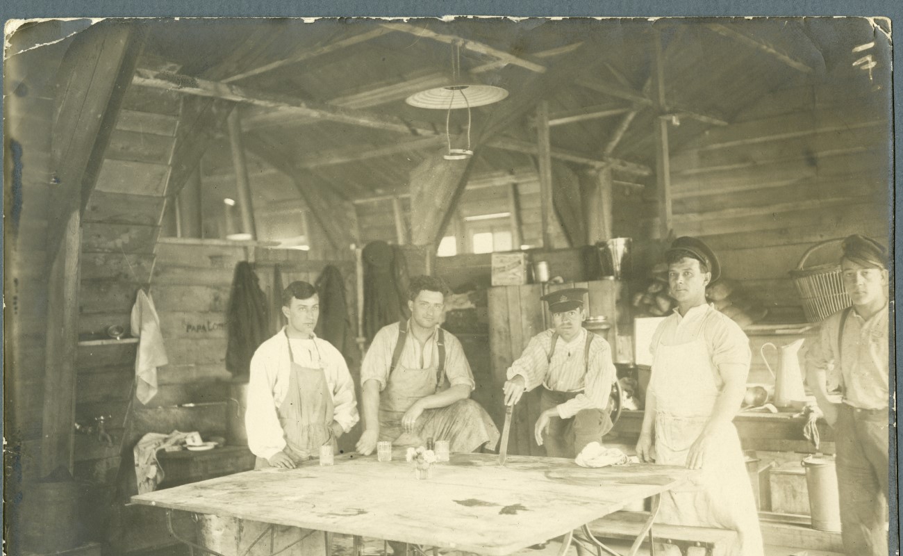Fem vitklädda sjukvårdare iförda förkläden står vid ett vitt bord i en timrad byggnad.