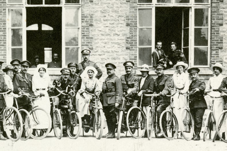 En grupp kvinnor och män i uniform och sjukvårdkläder står och poserar med cyklar utanför en tegelbyggnad med vidöppna fönster där fler personer skymtas.