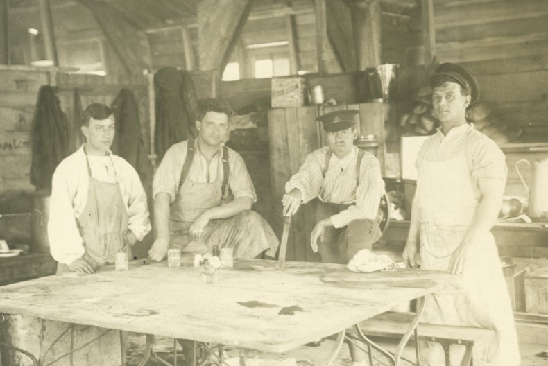 Fem vitklädda sjukvårdare iförda förkläden står vid ett vitt bord i en timrad byggnad.
