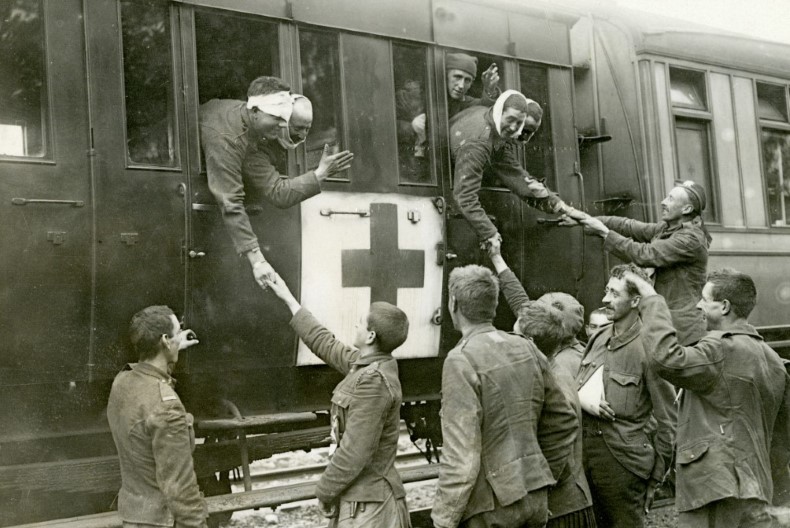 Skadade soldater hänger ut genom fönstret på ett tåg och tar farväl av sina kamrater ståendes på perrongen. Tåget är märkt med sjukvårdens kors.