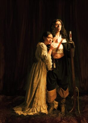 En person i vit böljande klänning lutar sig mot en annan person stödd på sitt gevär och klädd i uniform. Uniformsjackan är öppen och bröstet lindat med vitt tyg.