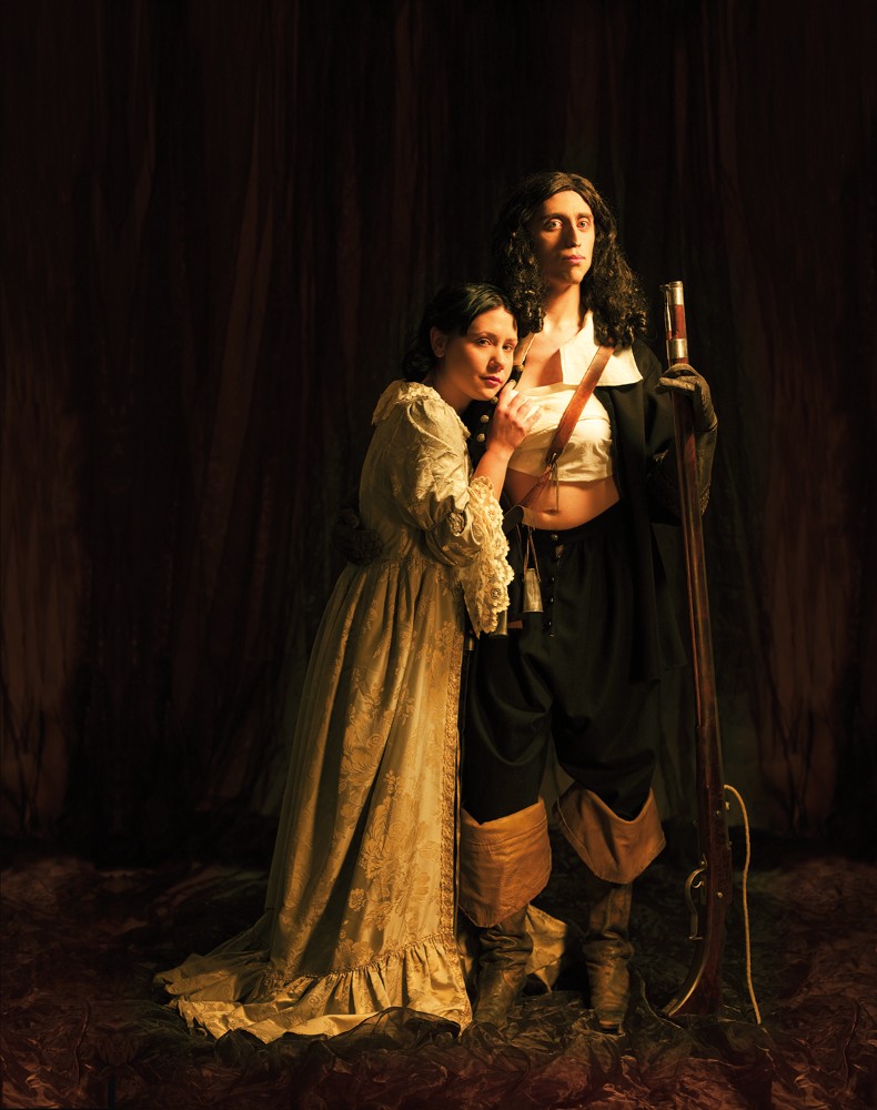 En person i vit böljande klänning lutar sig mot en annan person stödd på sitt gevär och klädd i uniform. Uniformsjackan är öppen och bröstet lindat med vitt tyg.