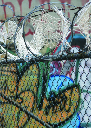 Framför en graffittimålad vägg står ett staket med taggtråd. Taggtråden är dekorerad med virkade smådukar som påminner om spindelnät där de sitter.