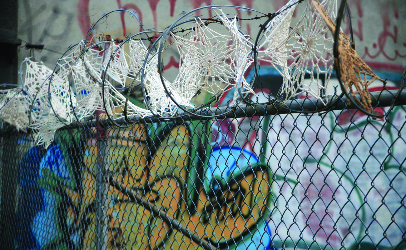 Framför en graffittimålad vägg står ett staket med taggtråd. Taggtråden är dekorerad med virkade smådukar som påminner om spindelnät där de sitter.