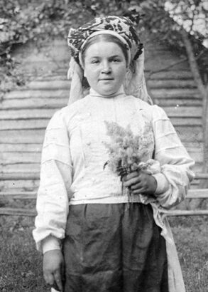 En person står framför ett timrat hus med grönska runtom, hon har en huvudbonad, vit blus och troligtvis en kjol på sig. I vänstra handen håller hon en blombukett.