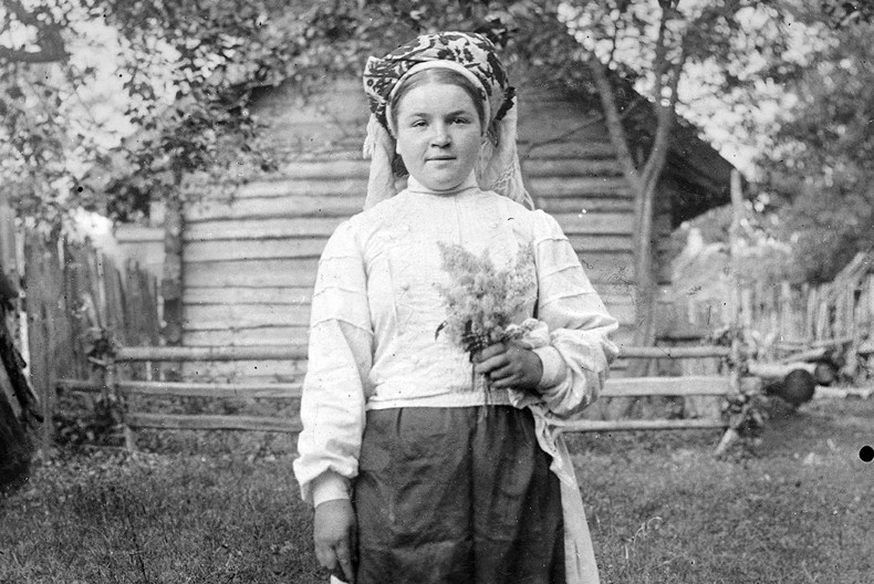 En person står framför ett timrat hus med grönska runtom, hon har en huvudbonad, vit blus och troligtvis en kjol på sig. I vänstra handen håller hon en blombukett.