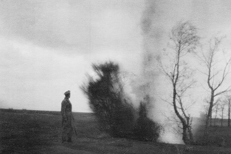 En militärklädd person med vapen i handen står på ett fält, med bara något enstaka träd, och ser bort i fjärran.