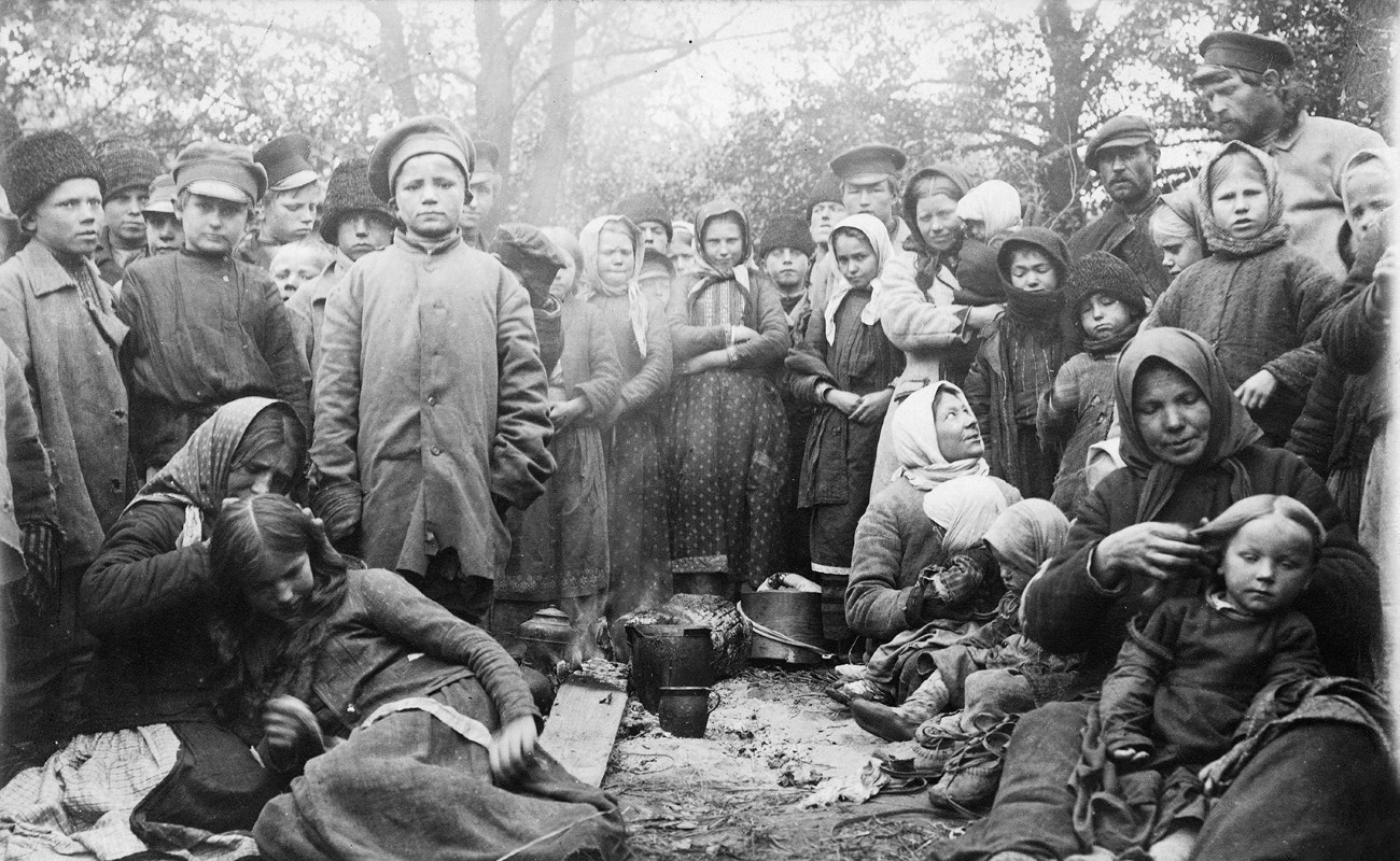En stor grupp människor, varav många barn, klädd i ytterkläder och ofta med något på huvudet, de flesta står upp, några sitter ner på marken. De flesta tittar in i kameran. I mitten på marken kastruller och kärl.
