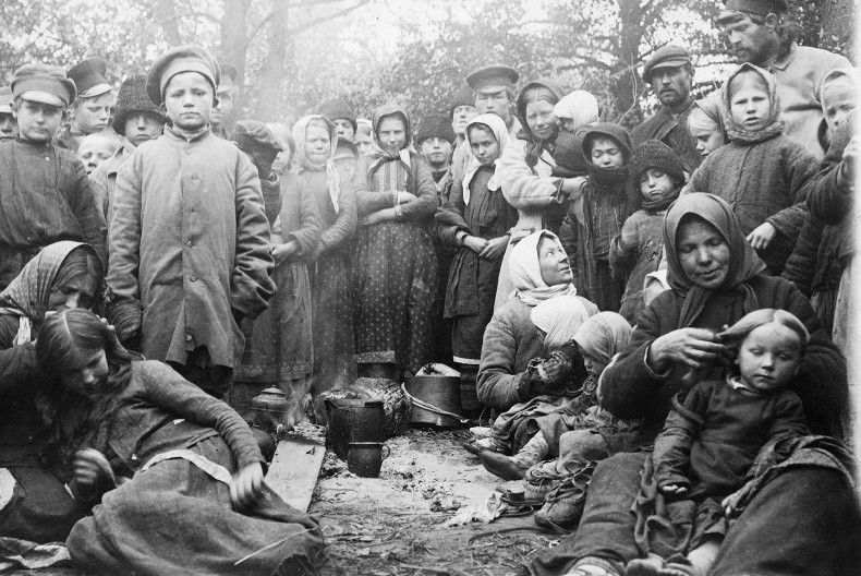 En stor grupp människor, varav många barn, klädd i ytterkläder och ofta med något på huvudet, de flesta står upp, några sitter ner på marken. De flesta tittar in i kameran. I mitten på marken kastruller och kärl.