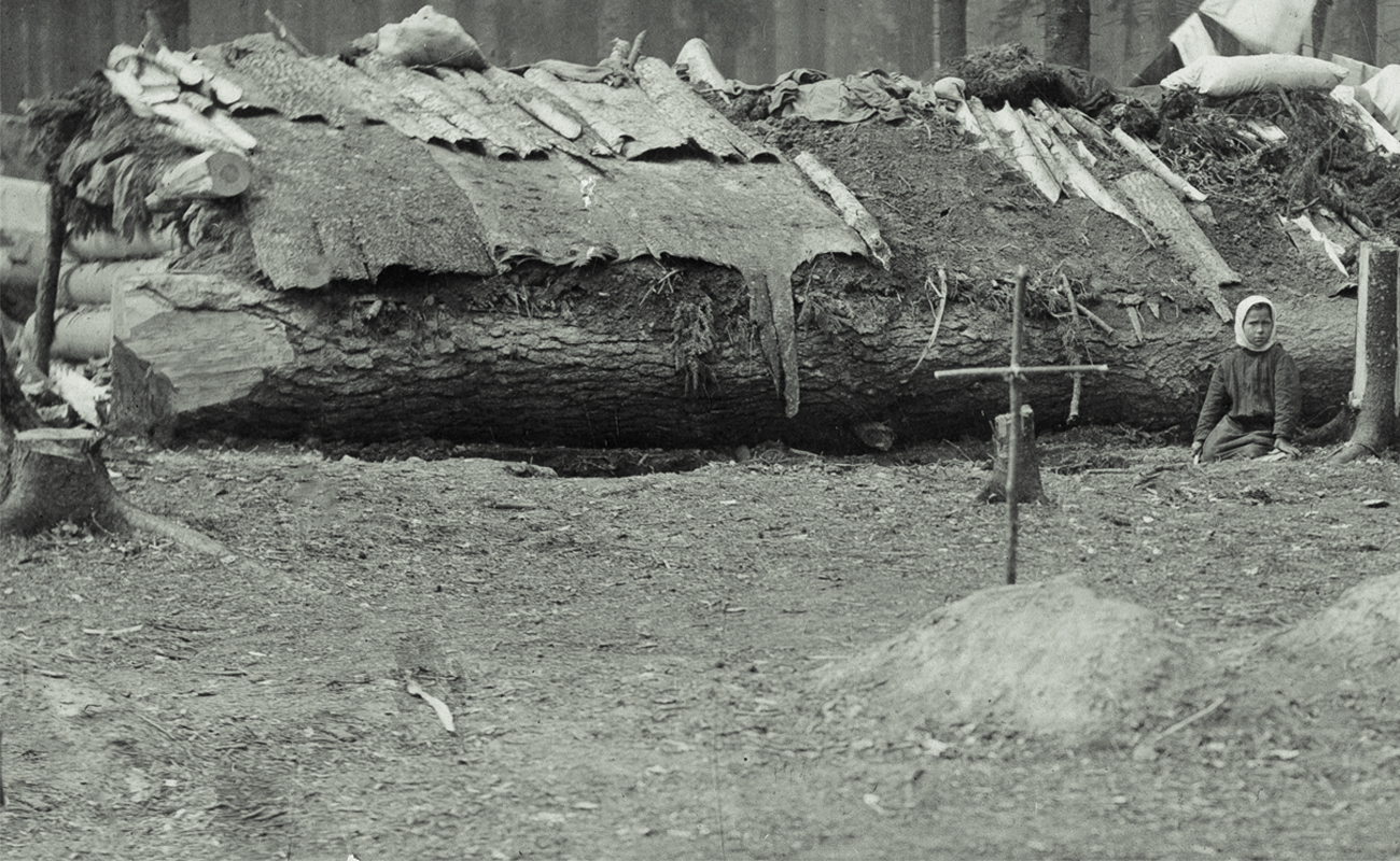 I förgrunden ett kors av pinnar nedstucket i jorden. I bakgrunden ett barn som sitter framför ett fällt träd som utgör basen i en stuga med lappat tak.