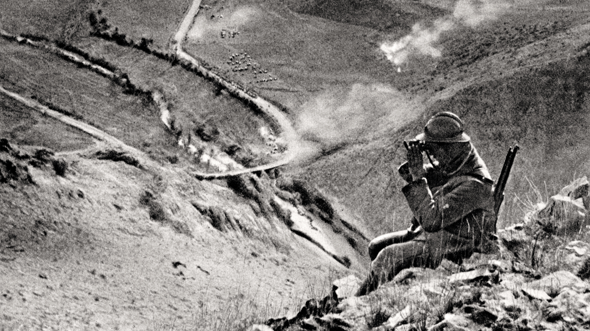 En man med vapen på ryggen sitter på en höjd i bergen och spanar med sin kikare. Längre bort syns en väg som snirklar sig uppåt.