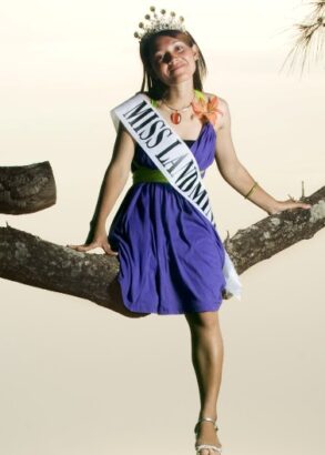 En person i lila klänning, tiara i håret och med ett satinband över bröstet "Miss landmine" sitter på en trädgren.