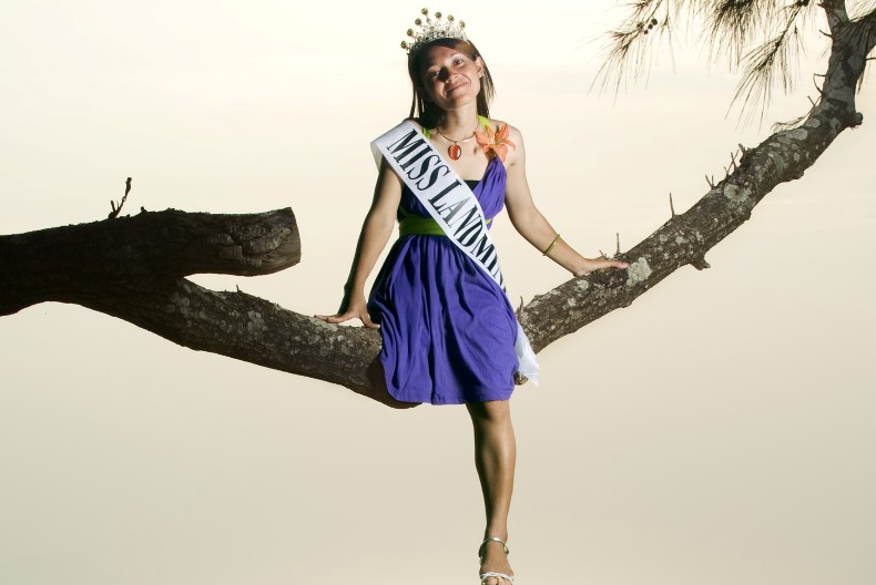 En person i lila klänning, tiara i håret och med ett satinband över bröstet "Miss landmine" sitter på en trädgren.