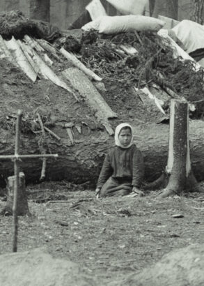 I förgrunden ett kors av pinnar nedstucket i jorden. I bakgrunden ett barn som sitter framför ett fällt träd som utgör basen i en stuga med lappat tak.